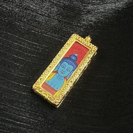 Close-up of the Medicine Buddha mini thangka pendant showing the hand-painted blue Buddha head, dark blue hair bun, red background, and ornate gold-tone raised border on the pendant frame.