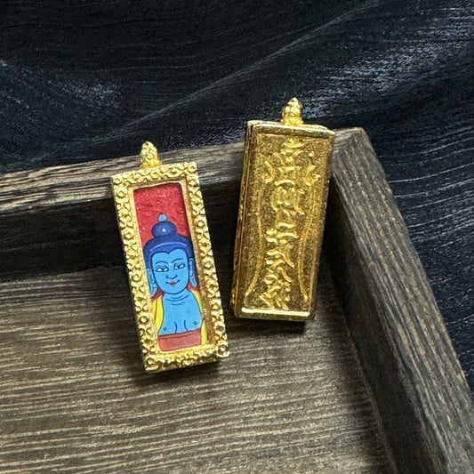 Front and back view of a Medicine Buddha mini thangka pendant, showing the hand-painted blue Buddha head on a red ground and the embossed script on the copper case, with an ornate gold-tone frame.