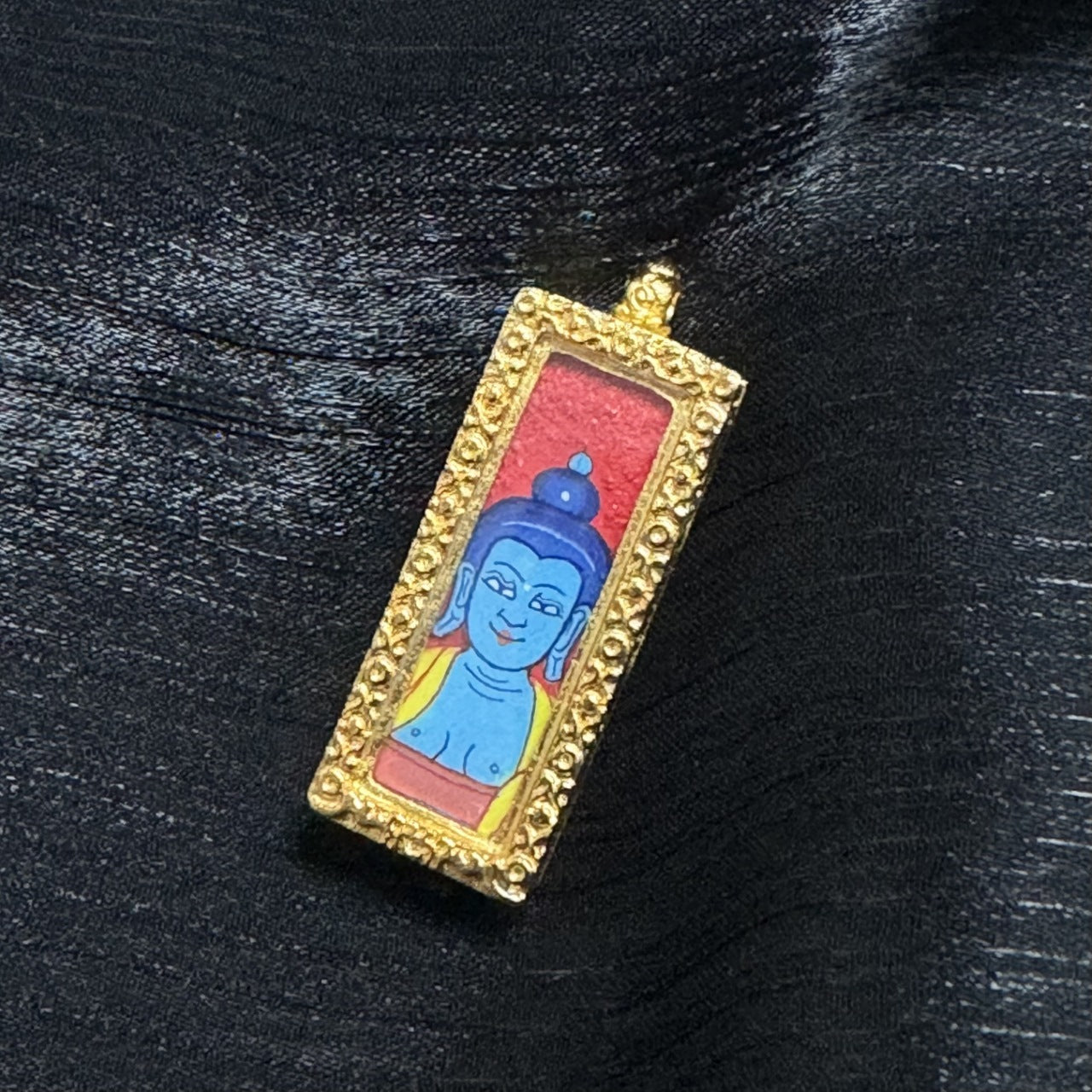 Front view of the Medicine Buddha mini thangka pendant showing the blue Buddha head painting on a red background, framed by an ornate raised gold-tone border and topped with a small hanging loop.