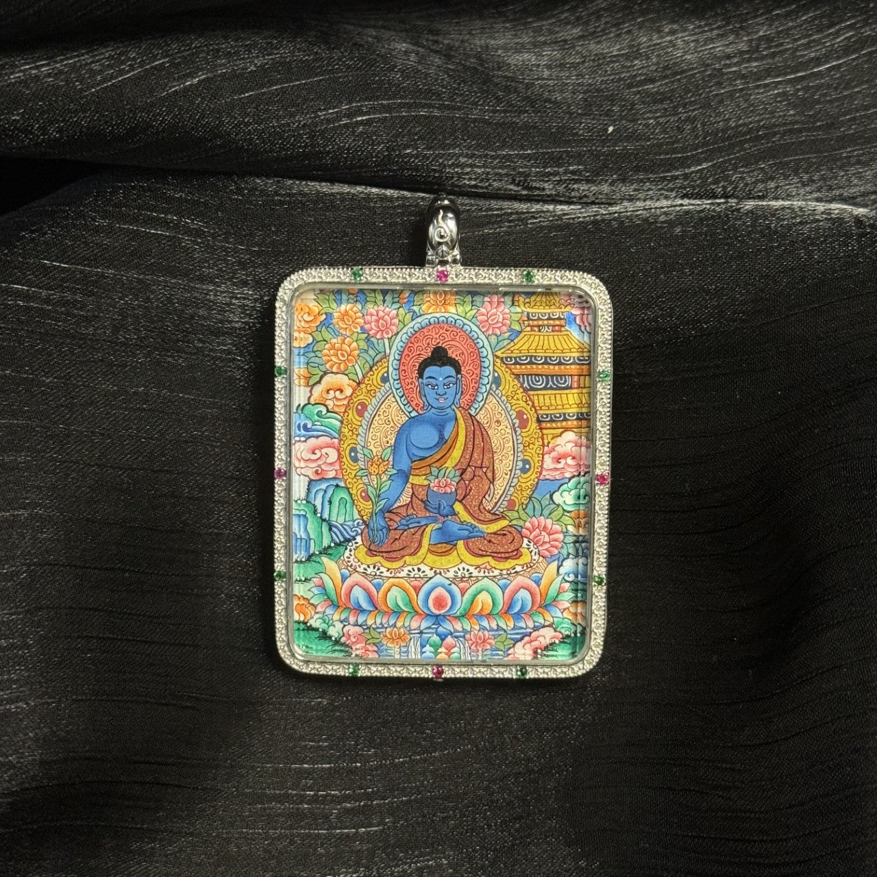 Hand-painted Tibetan Medicine Buddha miniature thangka pendant showing the seated Buddha on a lotus throne surrounded by traditional floral and architectural motifs.