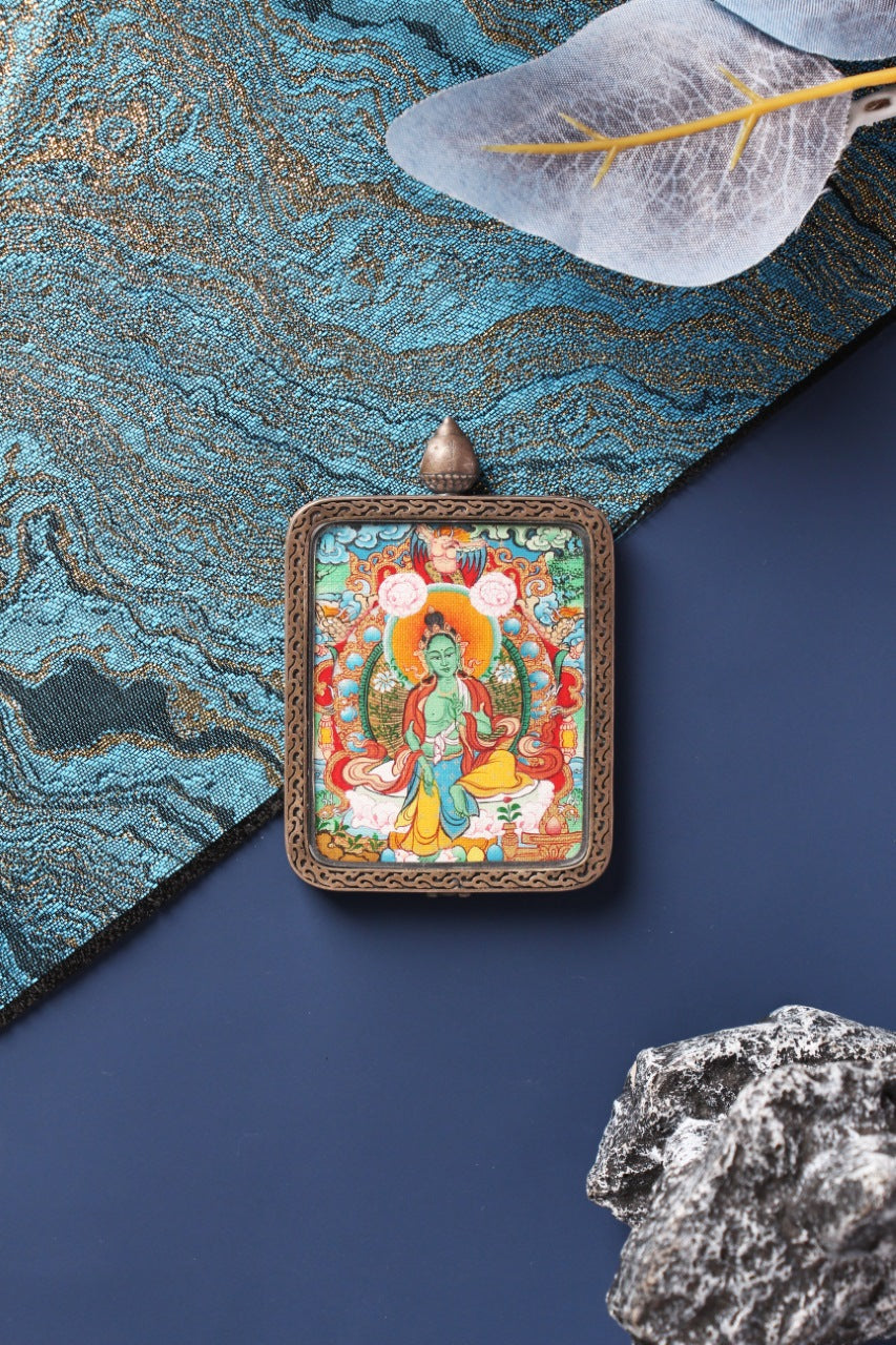 Centered front view of a Menri style Green Tara mini thangka pendant, Tibetan mineral pigment artwork set in a Dunhuang Nine-Color Deer silver case, photographed with blue-gold textile and stone accents.