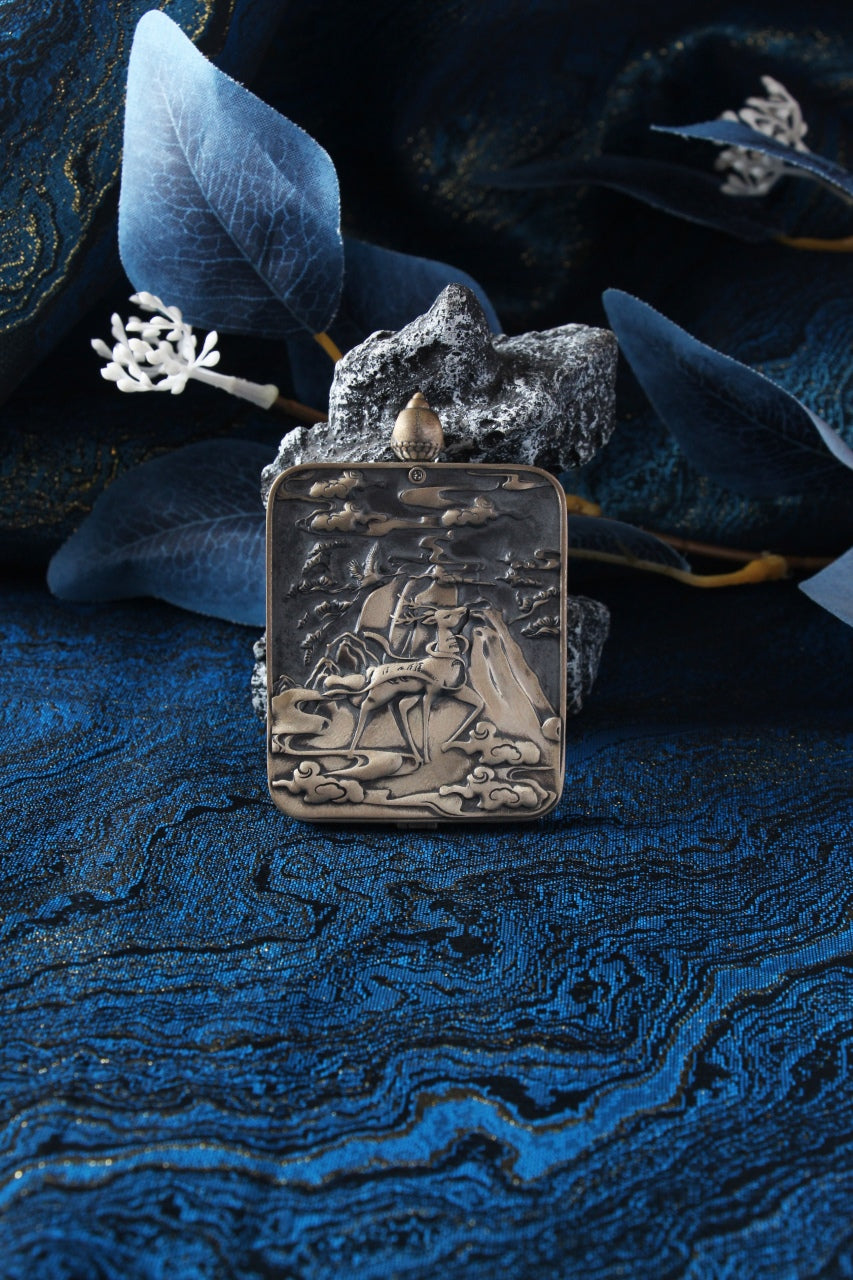 Back view of a Menri style mini thangka pendant in a Dunhuang Nine-Colored Deer silver case, photographed on blue fabric with rocks.