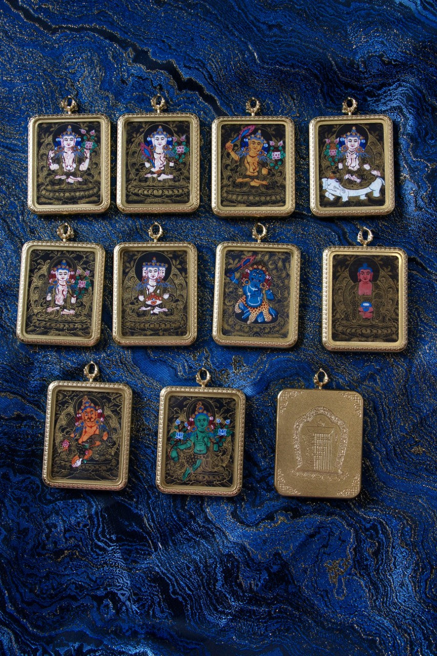 Assorted Nepalese black-gold mini thangka deity pendants displayed together on blue brocade fabric, including a Yellow Jambhala pendant in a gold-tone case