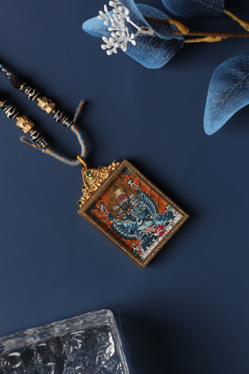 Nepal-style Yamantaka thangka pendant necklace with braided cord and beads, set in a jeweled double-sided titanium steel case on a dark blue flat-lay background.