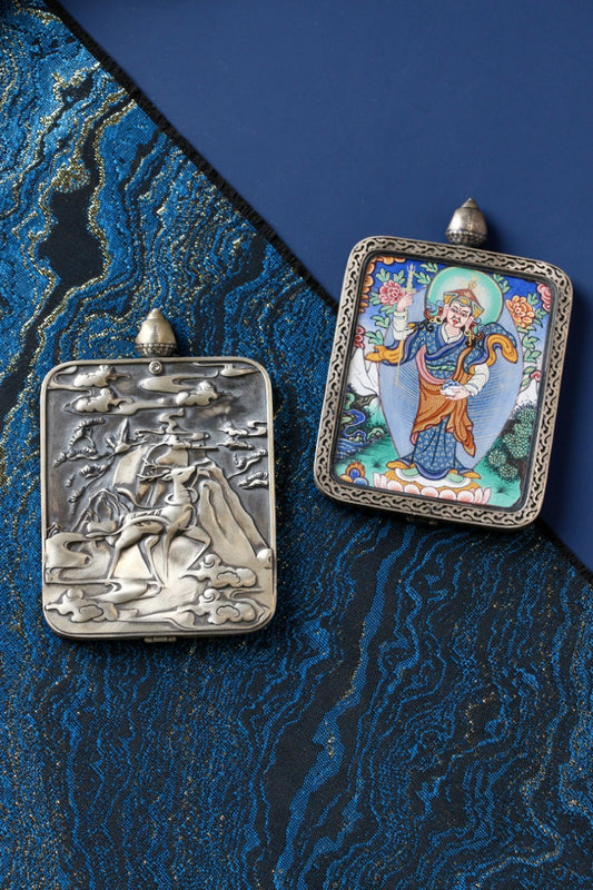 Padmasambhava (Guru Rinpoche) standing form mini thangka pendant shown beside a silver gawu case cover on blue textile background