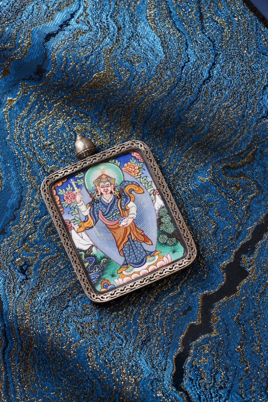 Padmasambhava standing form mini thangka pendant placed on blue brocade fabric, showing the hand-painted artwork inside a silver frame