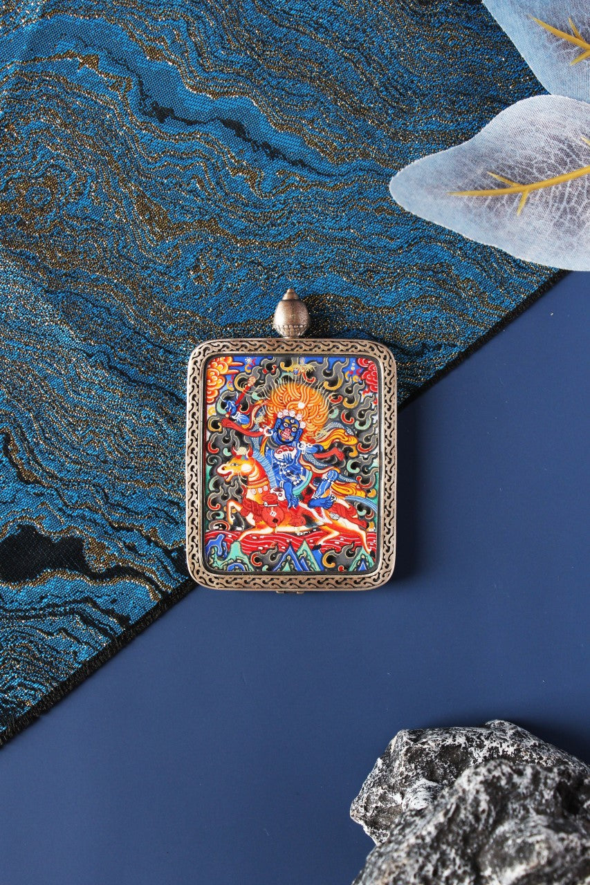 Flat lay of a Palden Lhamo protector deity mini thangka pendant with vibrant Rebgong painting and a 925 silver frame on blue brocade fabric background.