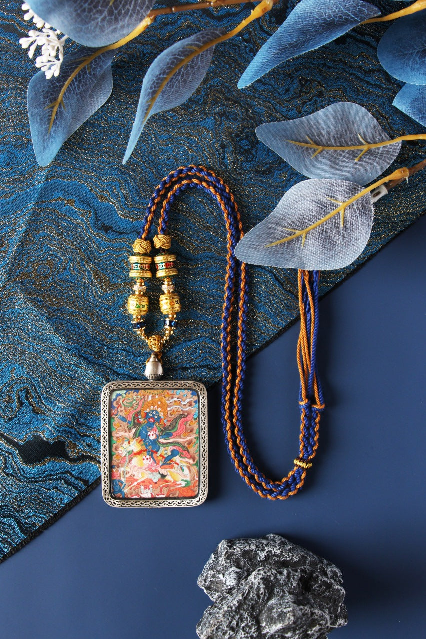 Full set display of a Palden Lhamo mini thangka pendant with braided necklace and tassel cords, showing the hand-painted artwork framed in a silver protective case on a blue brocade background with studio props