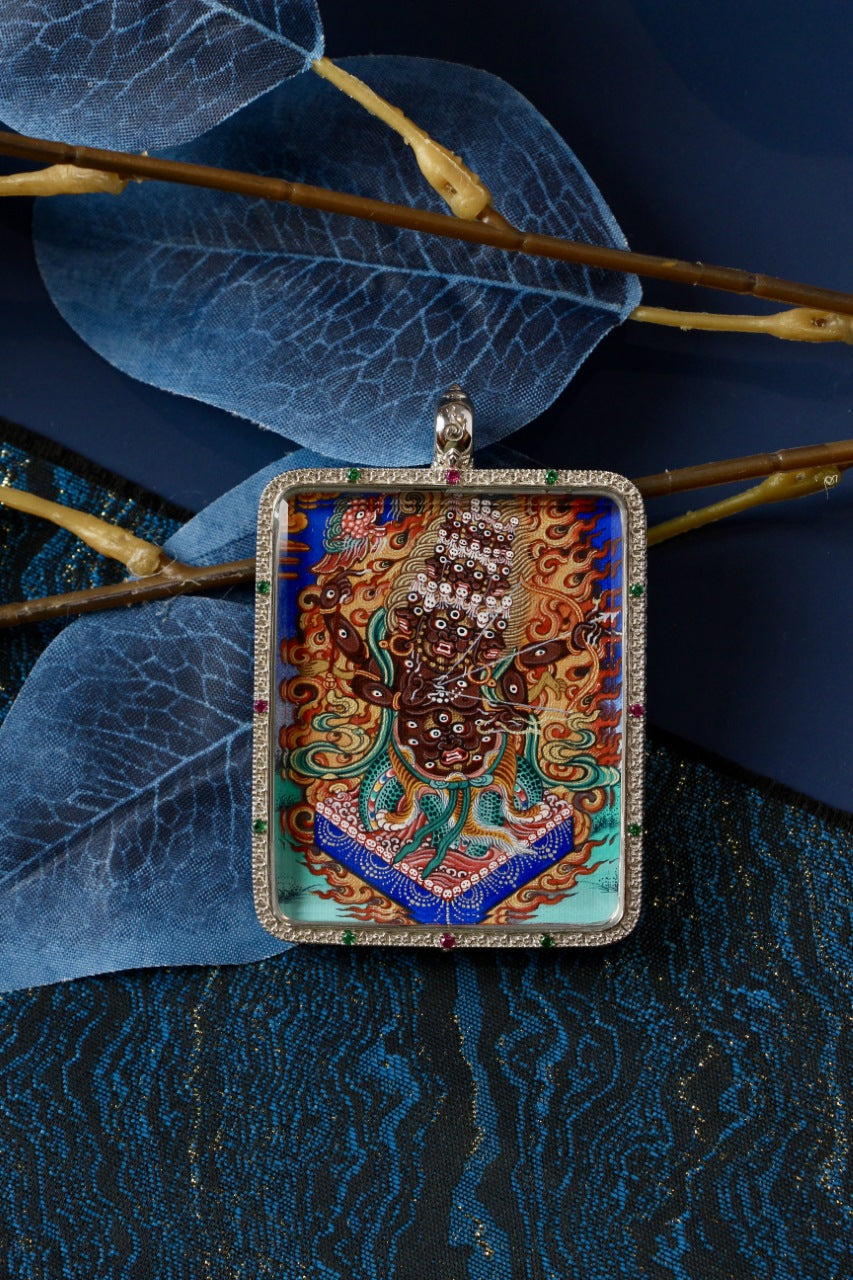 Front view of the Rahu (Rahula) hand-painted Rebgong thangka pendant in a 925 silver Gawu case, styled with translucent leaf props and blue fabric to emphasize the artwork’s color and detail.