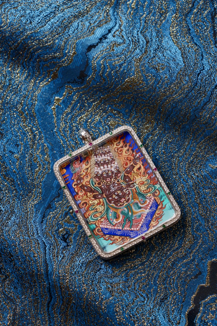 Angled flat-lay of a Rahu (Rahula) Rebgong thangka pendant in a 925 silver Gawu case, highlighting the vivid wrathful figure and flame motifs against a blue brocade surface.