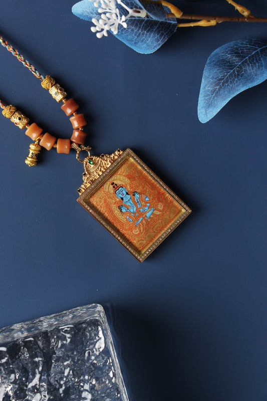 Angled view of Akashagarbha mini thangka pendant showing fine gold thangka linework, layered mineral pigment texture, gemstone-accented titanium steel frame, and braided cord connection with bead elements