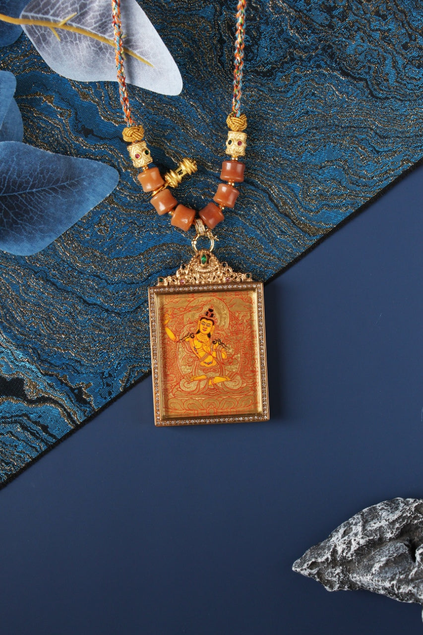 Angled view of Manjushri mini thangka pendant showing fine gold thangka linework, layered mineral pigment texture, gemstone-accented titanium steel frame, and braided cord connection