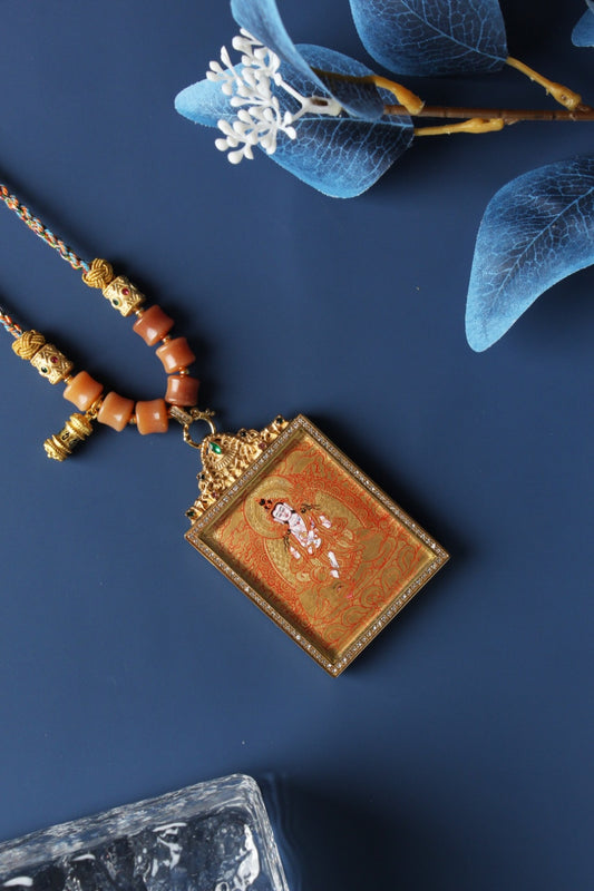 Angled view of Samantabhadra mini thangka pendant showing fine gold thangka linework, layered mineral pigment texture, gemstone-accented titanium steel frame, and braided cord connection