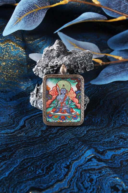 Close-up portrait of Akashagarbha Bodhisattva mini thangka pendant, fully hand-painted with mineral pigments, featuring vivid cloud motifs, a green halo, and fine linework, set in a Dunhuang Nine-Color Deer-inspired sterling silver case, photographed against textured blue brocade fabric with stone and layered translucent leaves.