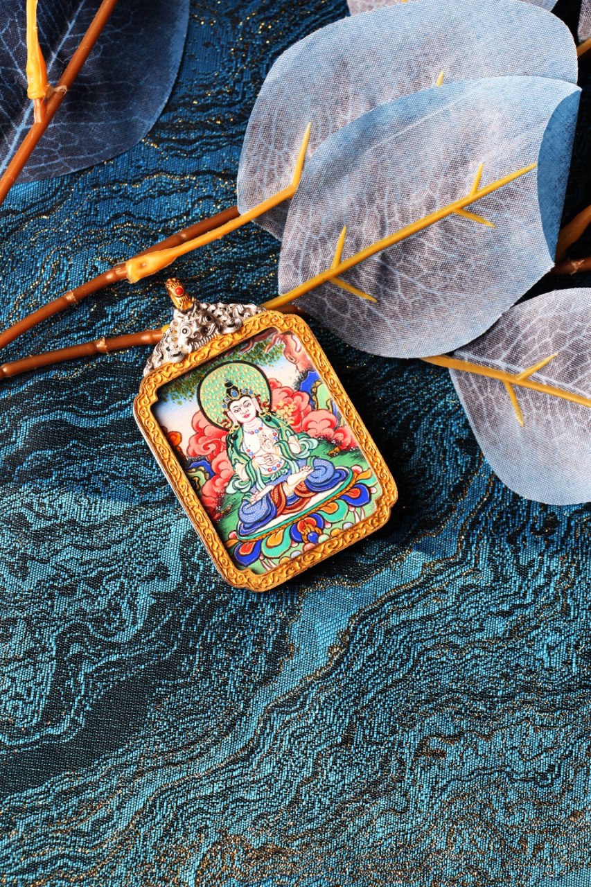 Angled close-up of Mahasthamaprapta Bodhisattva mini thangka pendant, hand-painted with mineral pigments and framed in an ornate silver-plated case with gold-tone border, photographed on blue patterned brocade with translucent leaves and warm-toned branches.