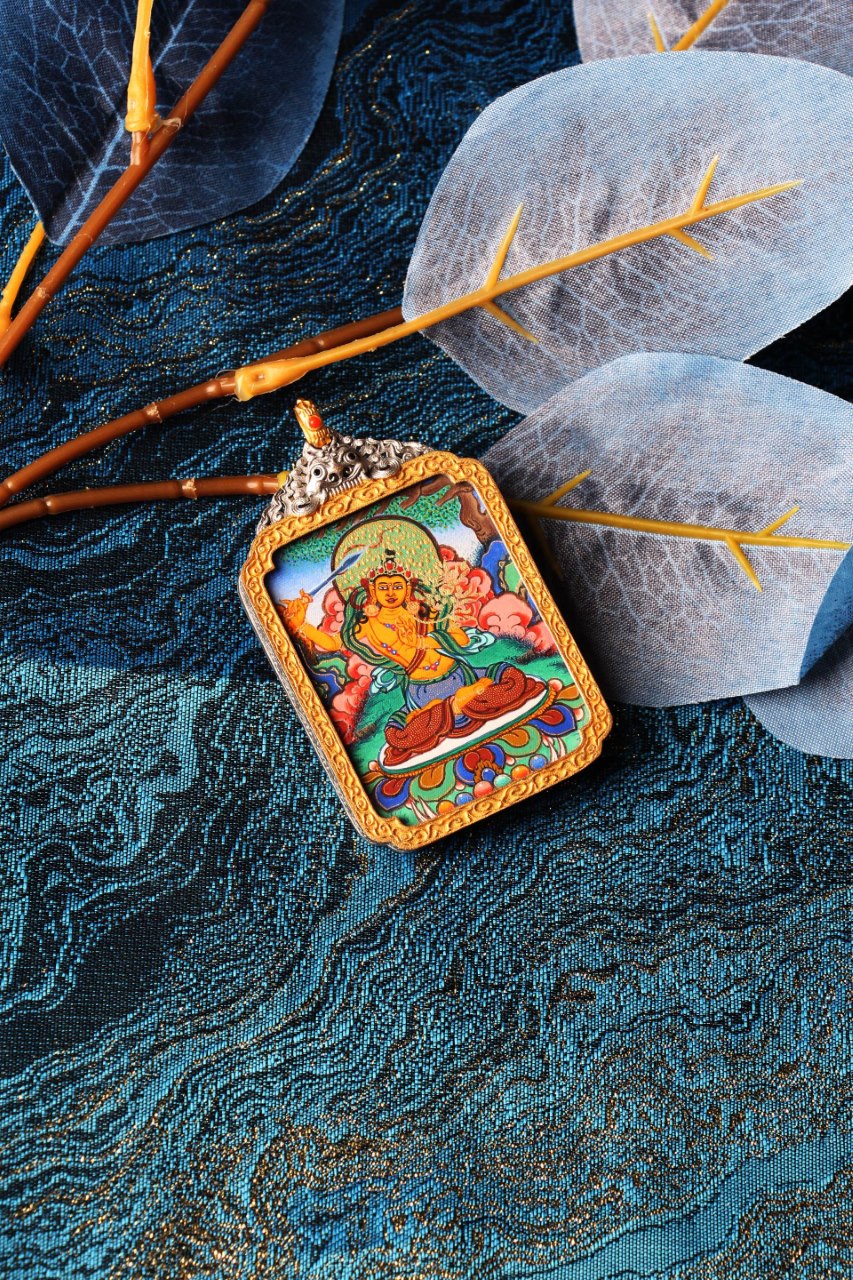 Angled close-up of Manjushri Bodhisattva mini thangka pendant, fully hand-painted with mineral pigments, highlighting vivid cloud motifs, green halo detailing, and fine linework, framed in a gold-tone border with an ornate silver-plated case top, photographed on blue patterned brocade with translucent leaf decor and warm-toned branch props.