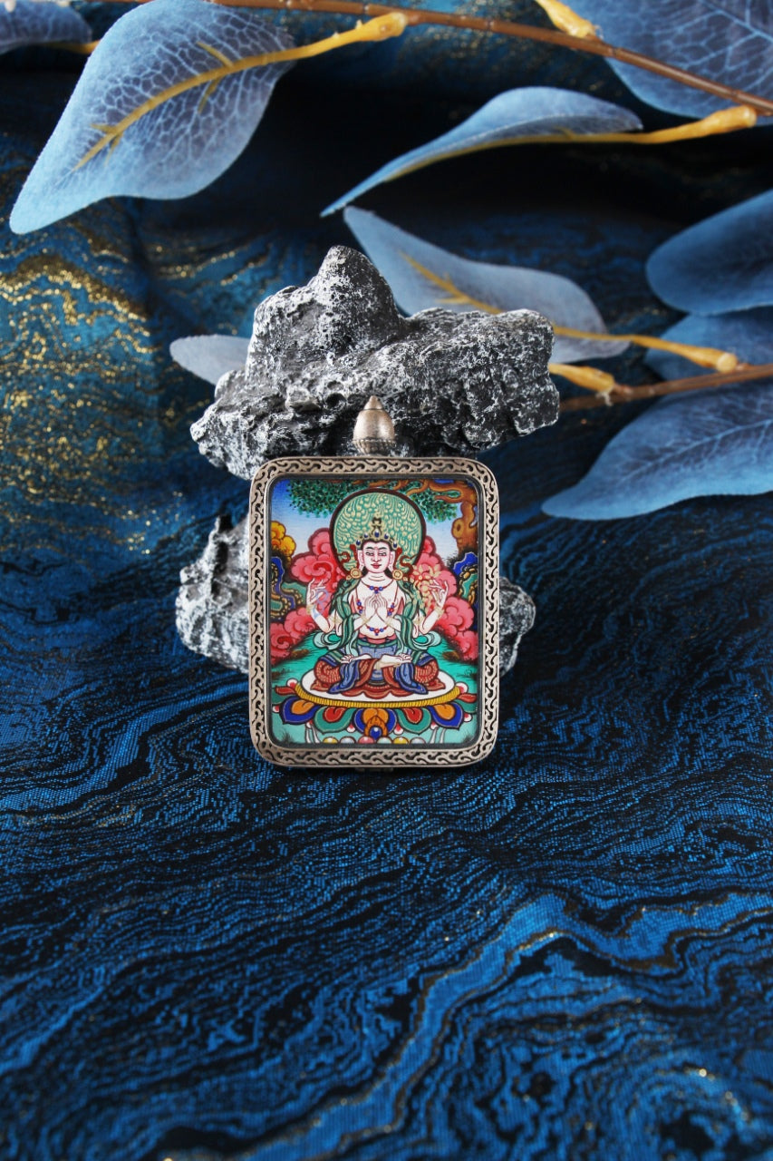 Close-up portrait of Four-Armed Avalokiteshvara mini thangka pendant, fully hand-painted with mineral pigments, highlighting fine linework, layered red cloud patterns, and detailed lotus base, set in a dark-toned sterling silver Dunhuang Nine-Color Deer case, photographed against textured blue brocade fabric with stone and layered translucent leaves.