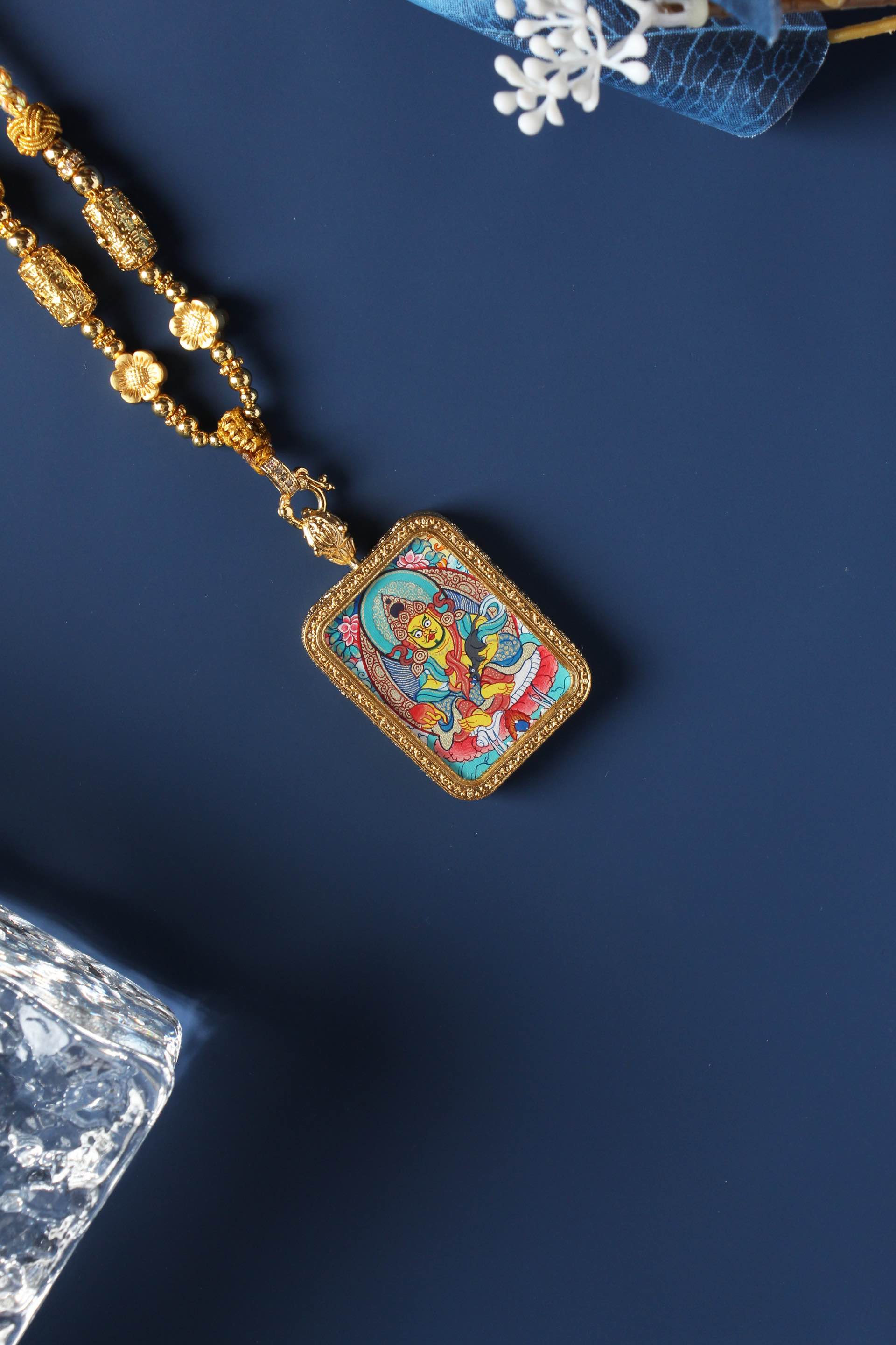 Angled view of Yellow Jambhala mini thangka pendant showing detailed iconography, mineral pigment layers, and gold-tone frame construction