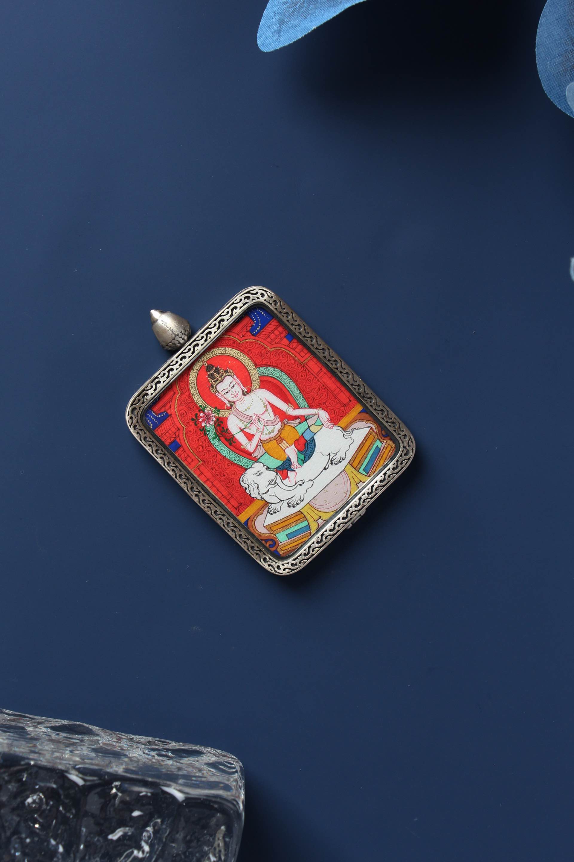 Flat lay of a hand-painted Samantabhadra Bodhisattva mini thangka pendant in a sterling silver case on a dark blue background