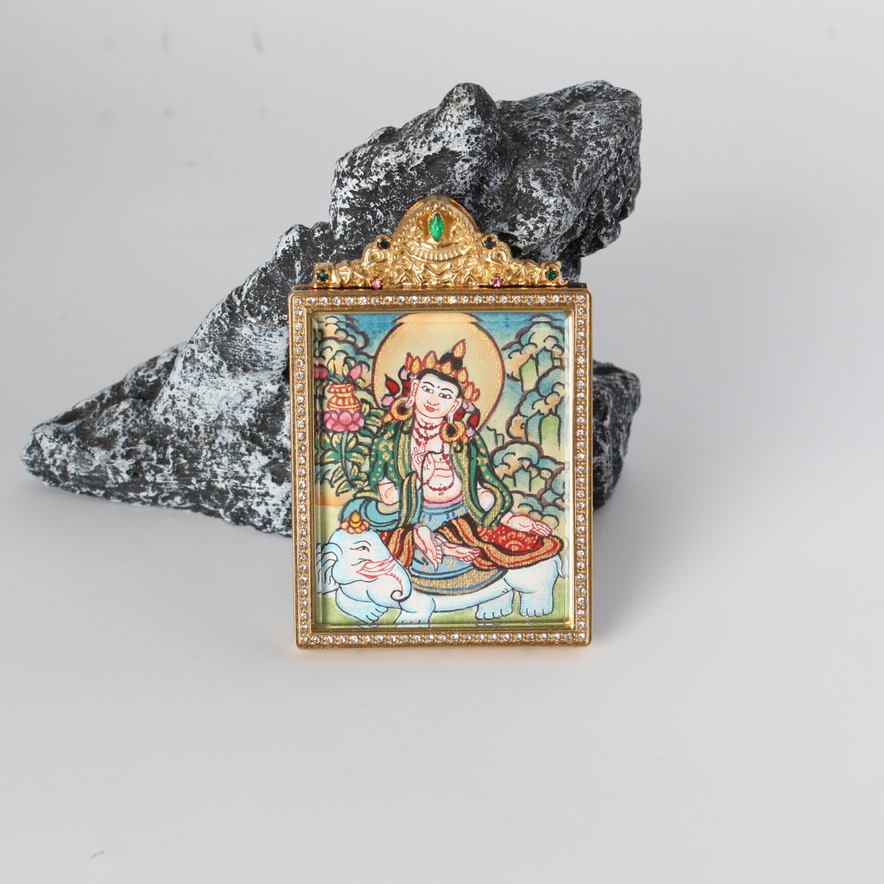 Front view of Samantabhadra Bodhisattva mini thangka pendant showing the hand-painted bodhisattva seated on a white elephant with halo and floral details, gold-tone rectangular frame with crystal border, and ornate crown top with a green gemstone, photographed on a rock prop.
