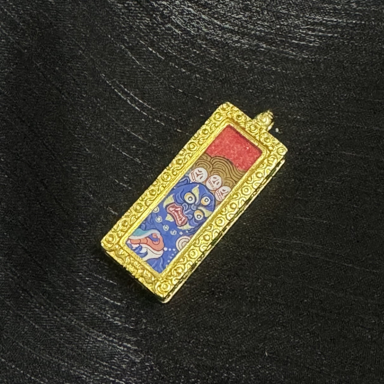 Angled view of a Six-Armed Mahakala mini thangka pendant showing the slim rectangular form, ornate raised frame texture, and the hand-painted blue deity face against a red upper background.