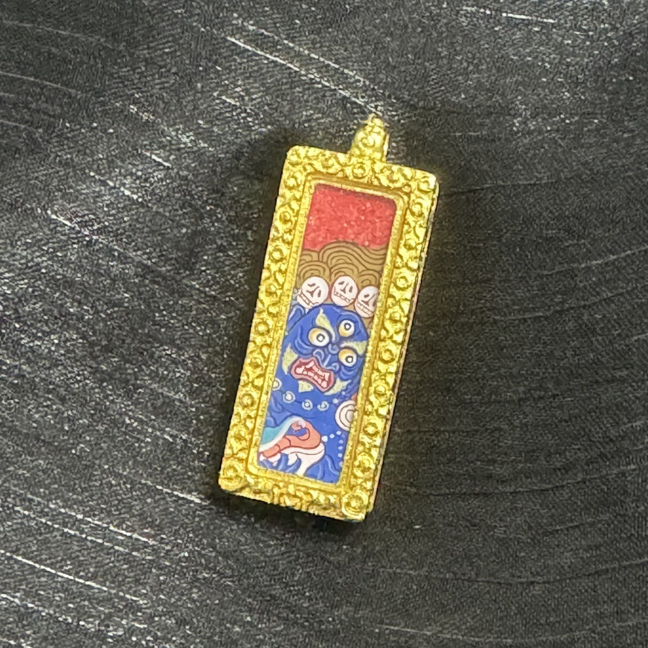 Front view of a Six-Armed Mahakala mini thangka pendant highlighting the red upper background, patterned golden section, multi-eye blue face, and the gold-tone raised border surrounding the painting within the copper case.