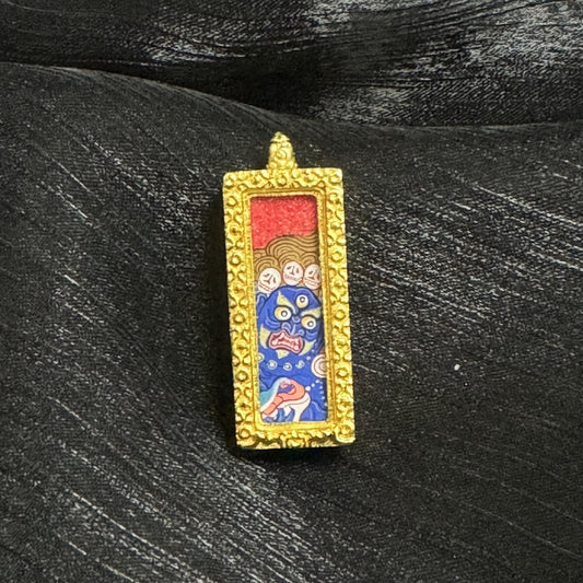 Front view of a Six-Armed Mahakala mini thangka pendant featuring a blue wrathful face with multiple eyes, expressive mouth details, red-and-gold upper section, and a gold-tone raised border around the pendant frame.