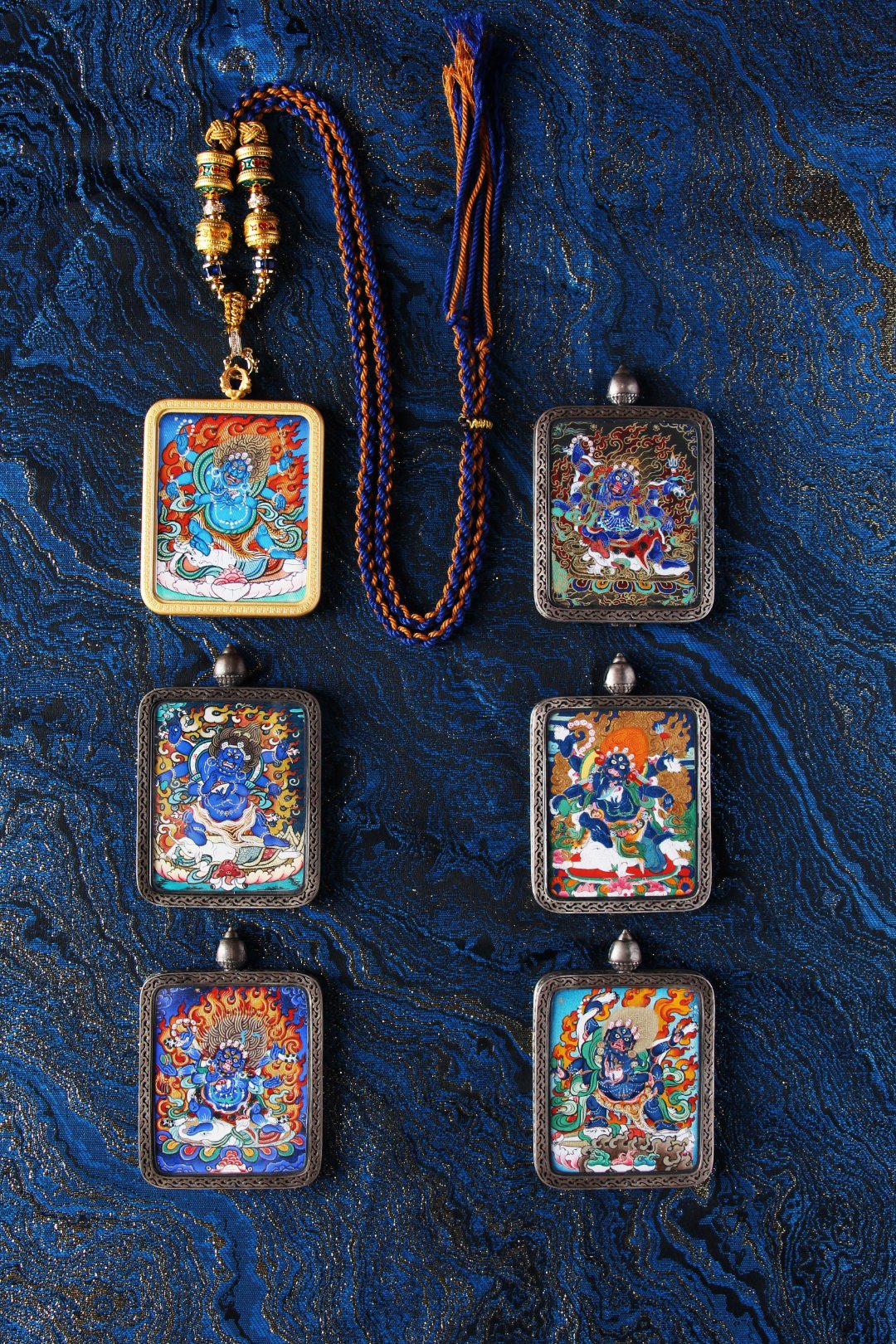 Group display of Six-Armed Mahakala mini thangka pendants with hand-painted mineral pigment artwork, shown in copper cases with braided blue-and-orange cord on a textured blue fabric background.
