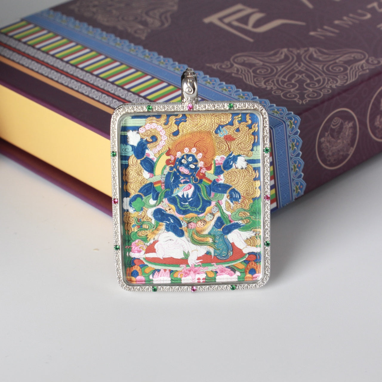 Straight-on view of the Six-Armed Mahakala mini thangka pendant highlighting textured mineral pigment application, gold-toned background details, and the silver frame with green and pink stone accents.