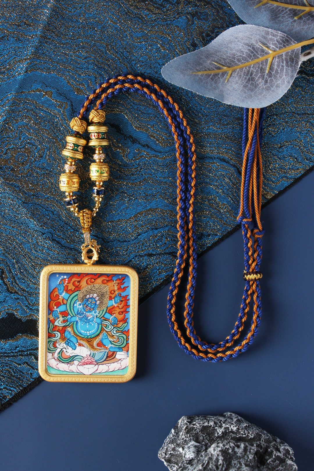 Six-Armed Mahakala mini thangka pendant shown with braided blue-and-orange cord and decorative bead section, highlighting the pendant’s front painting and metal case edge on a blue patterned fabric setting.