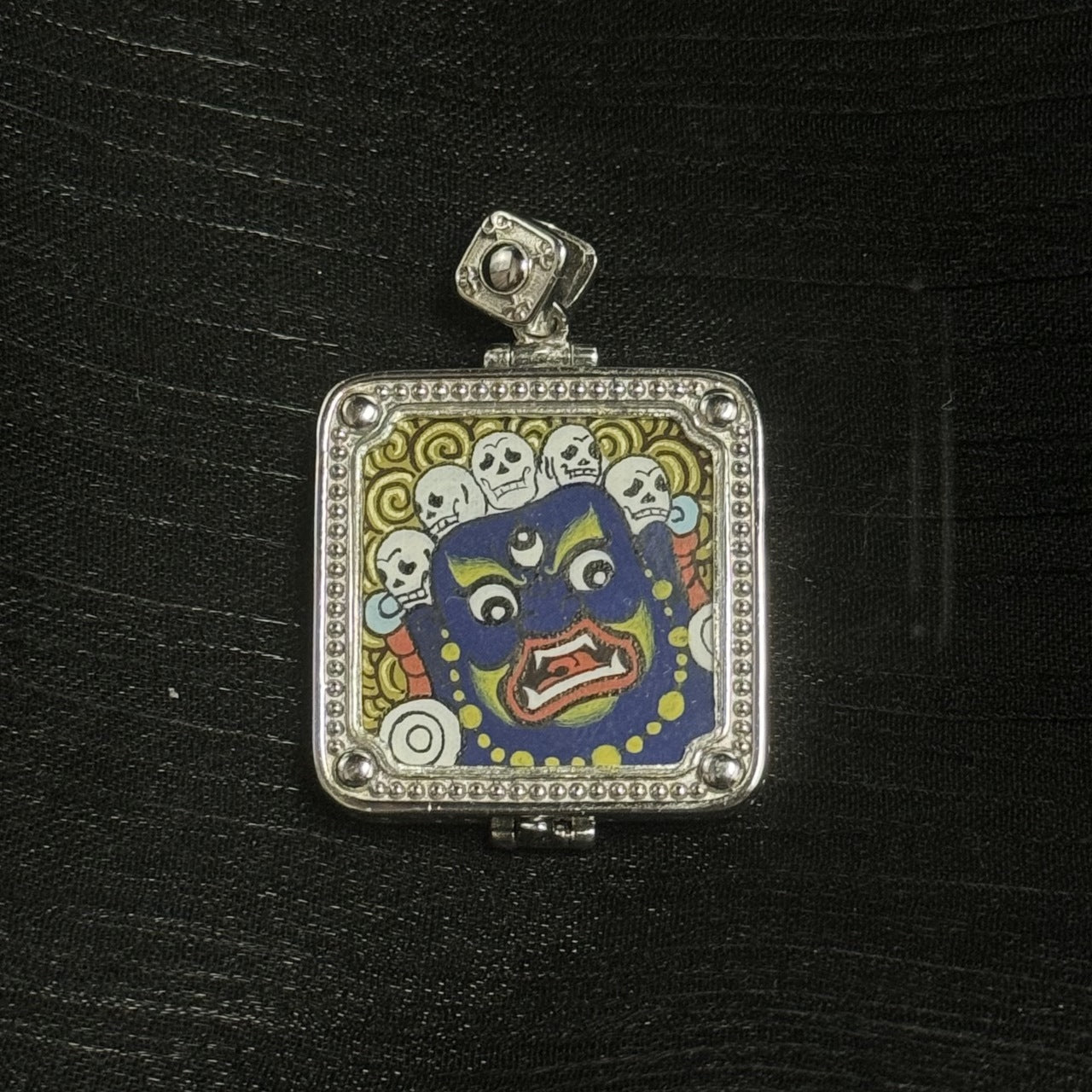 Close-up of wrathful Mahakala face painting with skull crown and swirling background patterns, framed in a square 925 silver electroplated gawu case with beaded border and corner studs