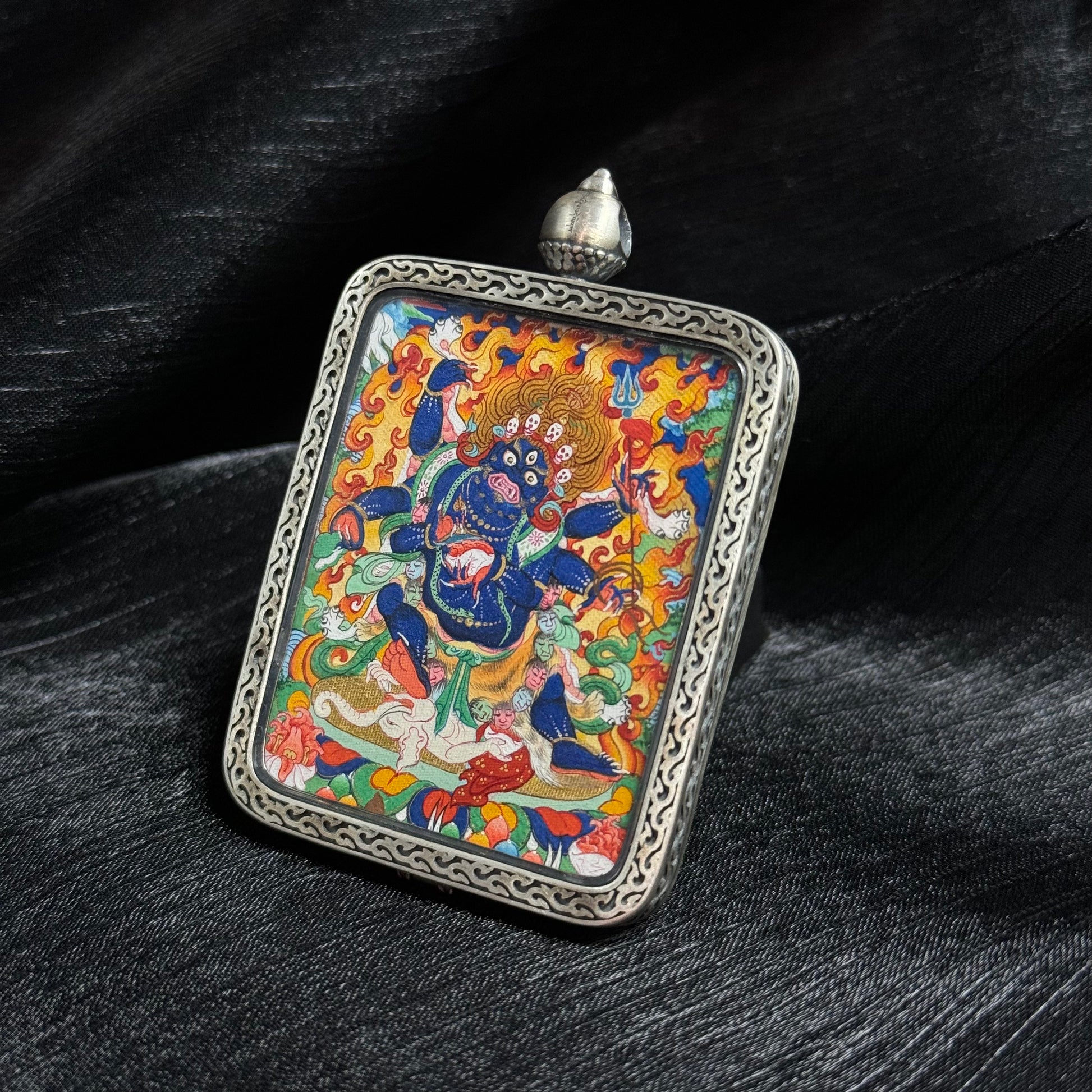 Angled view of Six-Armed Mahakala thangka pendant highlighting detailed mineral pigment painting, vivid flames and traditional Tibetan deity iconography inside a decorative silver frame