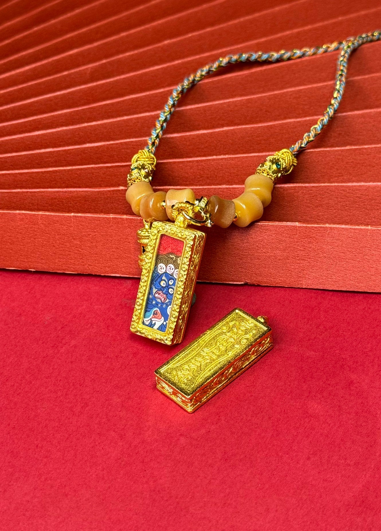 Angled view of Six-Armed Mahakala miniature thangka pendant showing cinnabar red background, blue mineral pigment face, and ornate copper pendant frame