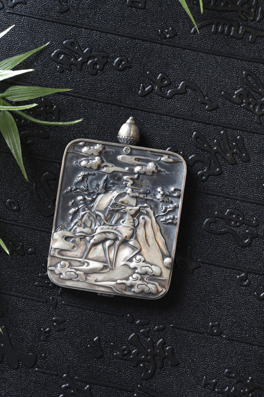 Back side of the sterling silver gawu pendant case with a raised landscape-and-deer engraving, photographed on a dark textured background.