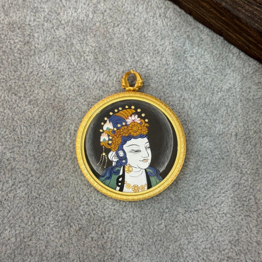 Close portrait view of a hand-painted Sudhana Avalokiteshvara mini thangka pendant, showing mineral pigment details, blue hair ornament, golden crown motifs, and a gold-tone round rim.
