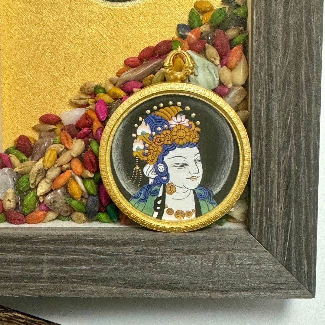 Sudhana Avalokiteshvara mini thangka pendant placed in a wooden tray, highlighting the round metal rim, dark halo background, and crisp fine-line painting around the face and crown.