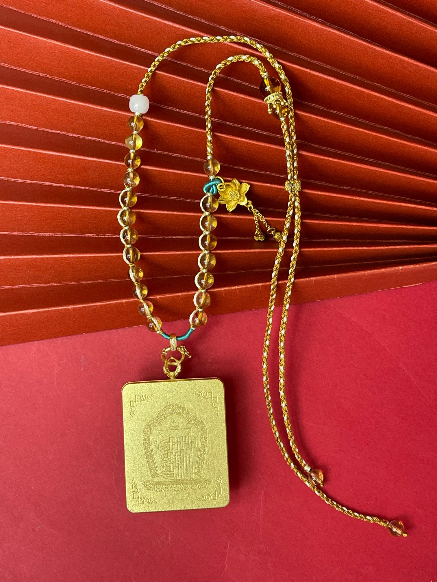 Close-up of the back side of a Thangka amulet pendant necklace displayed on a red background, featuring a rectangular pendant engraved with traditional religious symbols, paired with amber beads, decorative spacer beads, and a braided cord.