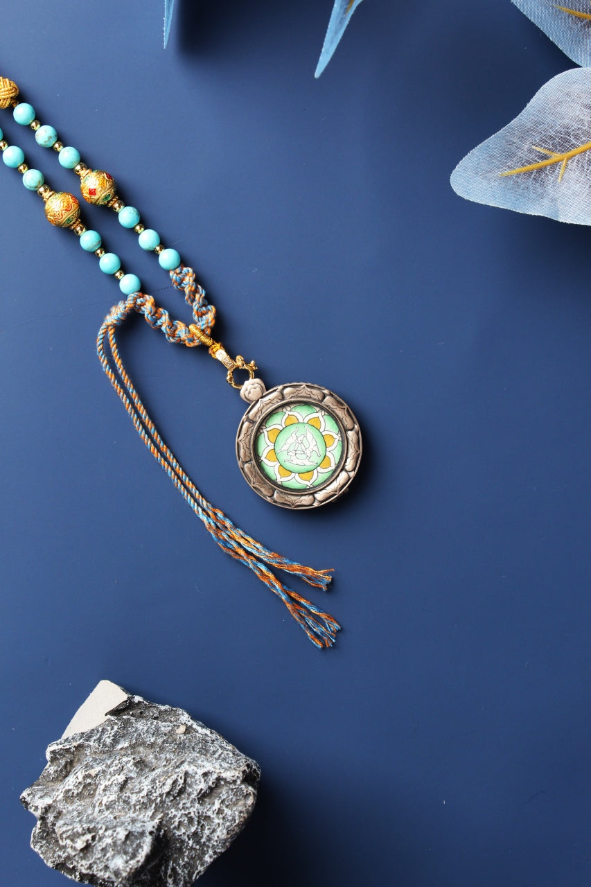 Full flat-lay photo of a round Three Hares mini thangka pendant in a 925 silver case, shown with a long braided cord, blue bead segments, tassel ends, and gold-tone decorative beads on a dark blue background.