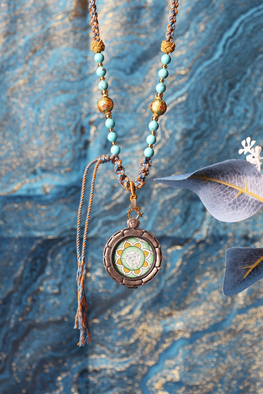 Vertical display photo of the Three Hares hand-painted mini thangka pendant in a round 925 silver case, showing the braided cord, turquoise-colored beads, and gold-tone bead accents against a textured blue backdrop.