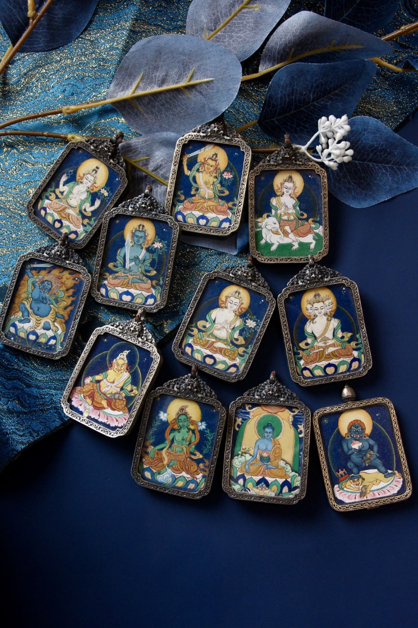 Assorted vintage-silver gawu pendants featuring hand-painted Tibetan mini thangka artworks on a blue textile background
