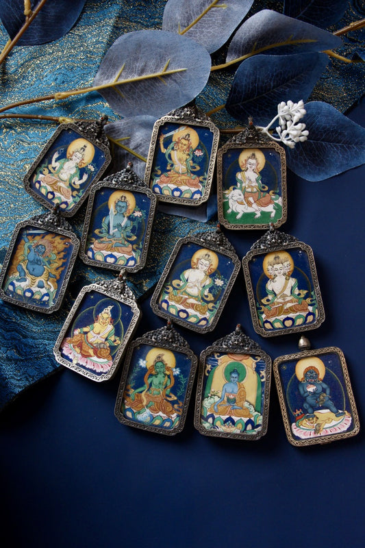 Assorted vintage-silver gawu pendants featuring hand-painted Tibetan mini thangka artworks on a blue textile background