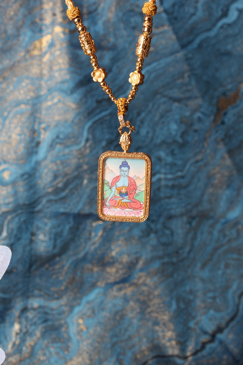 Hanging Bhaisajyaguru Buddha thangka pendant necklace in a gold-tone rectangular frame on a textured blue backdrop.