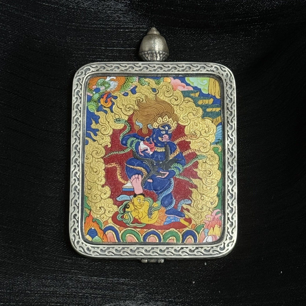 Detailed view of a Tibetan Black Jambhala mini thangka painting mounted in a silver gawu pendant case, highlighting the deity’s blue body, fiery golden halo, multicolored lotus base, and traditional Himalayan painting style.