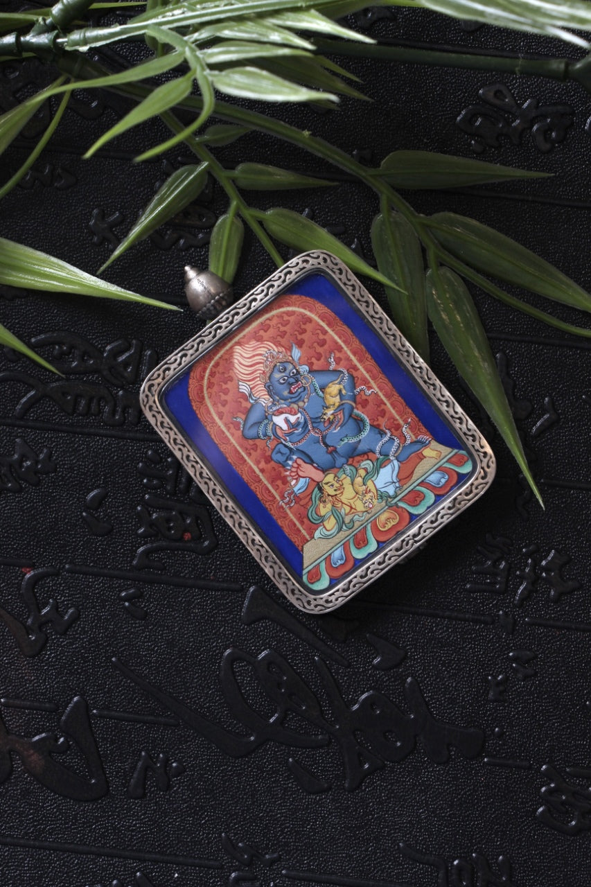 Angled product photo of a Tibetan Black Jambhala miniature thangka pendant in a silver amulet case, styled with bamboo leaves on a dark textured background.