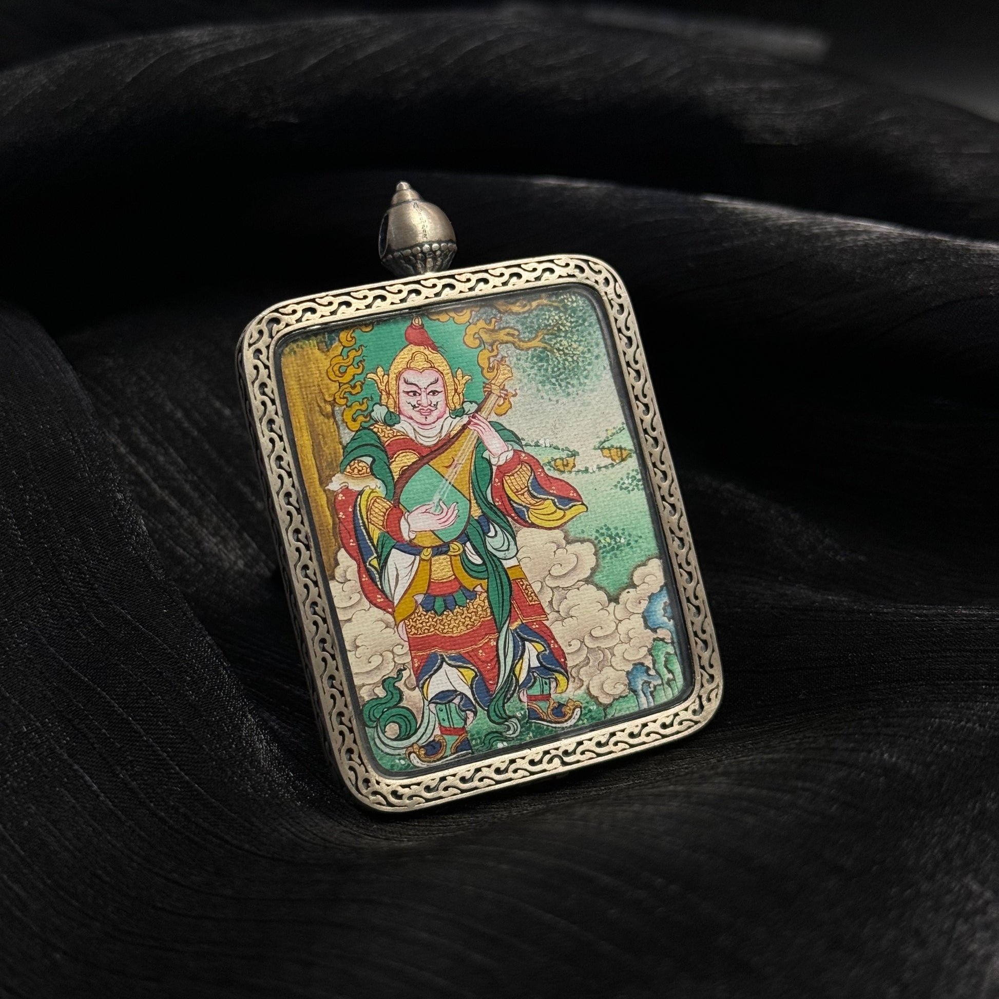 Close angled display of Dhṛtarāṣṭra mini thangka pendant highlighting mineral pigment colors, guardian king armor details, pipa instrument, and decorative sterling silver gawu-style pendant frame.