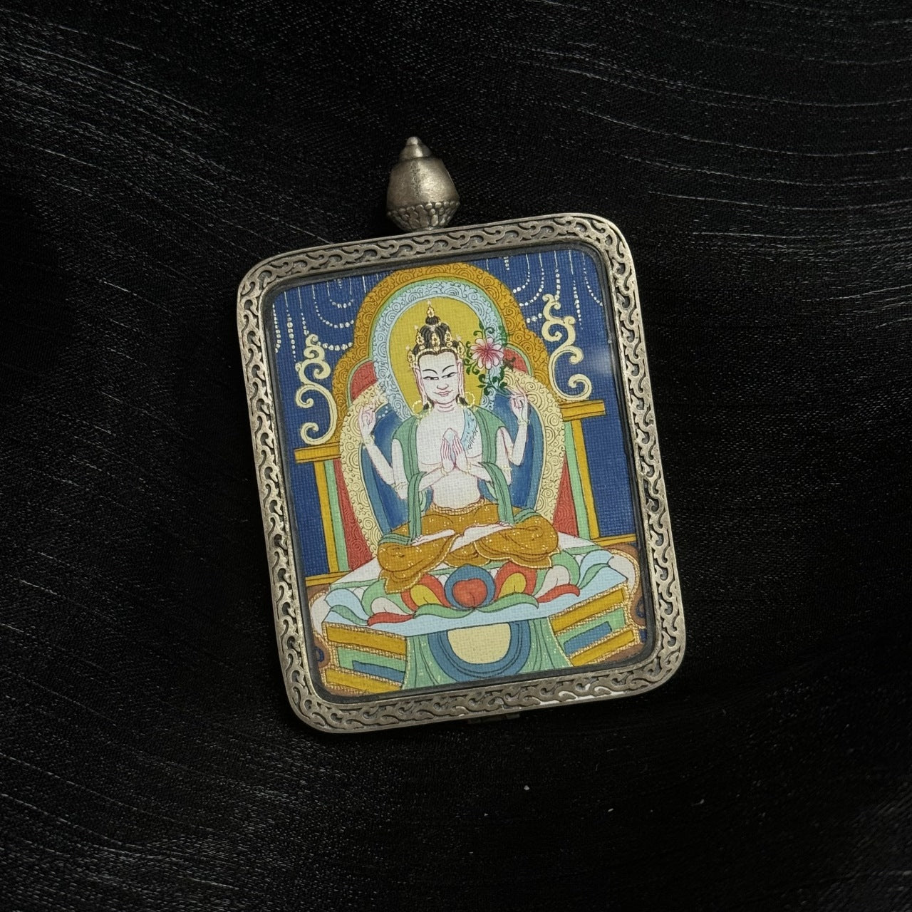 Four-Armed Avalokiteshvara thangka painting showing the bodhisattva of compassion with prayer gesture and lotus, framed in a decorative silver gawu pendant