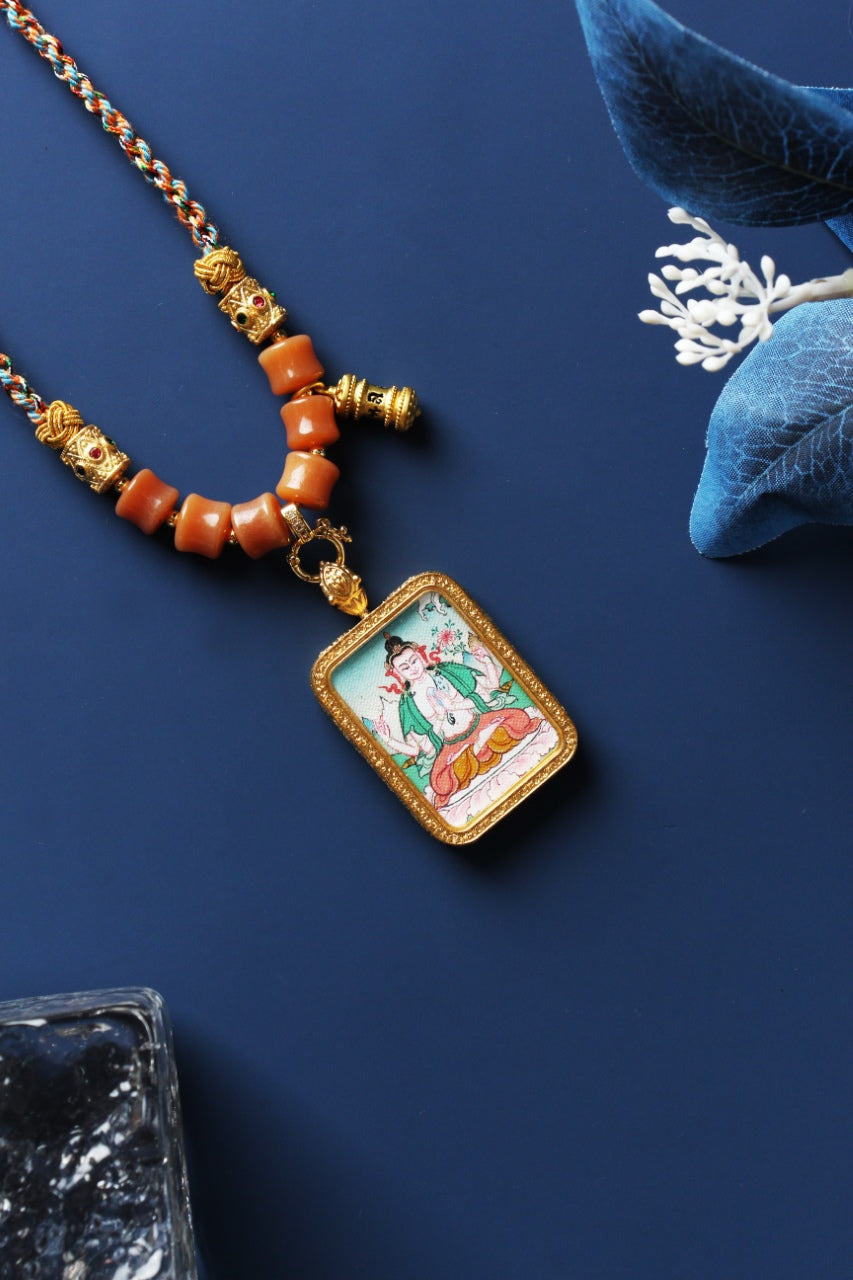 Tibetan Four-Armed Avalokiteshvara mini thangka pendant photographed on a blue background, featuring a rectangular metal frame, fine painted lines, and braided cord with bead sections