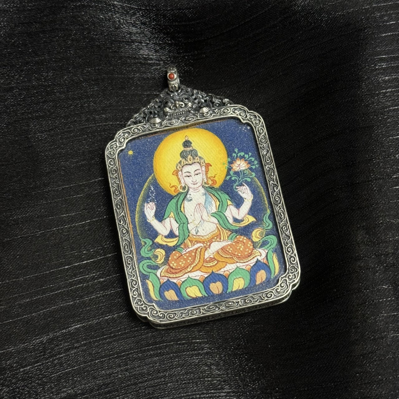 Front view of Four-Armed Avalokiteshvara mini thangka pendant showing golden halo, four-arm iconography, lotus seat, and layered mineral pigment background.