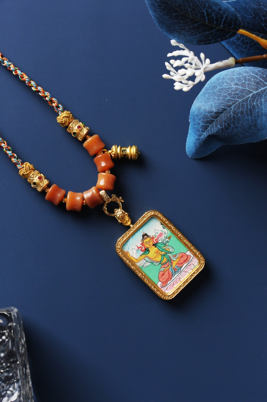 Tibetan Manjushri mini thangka pendant shown with braided cord necklace, cylindrical beads, decorative metal spacers, and the rectangular pendant frame
