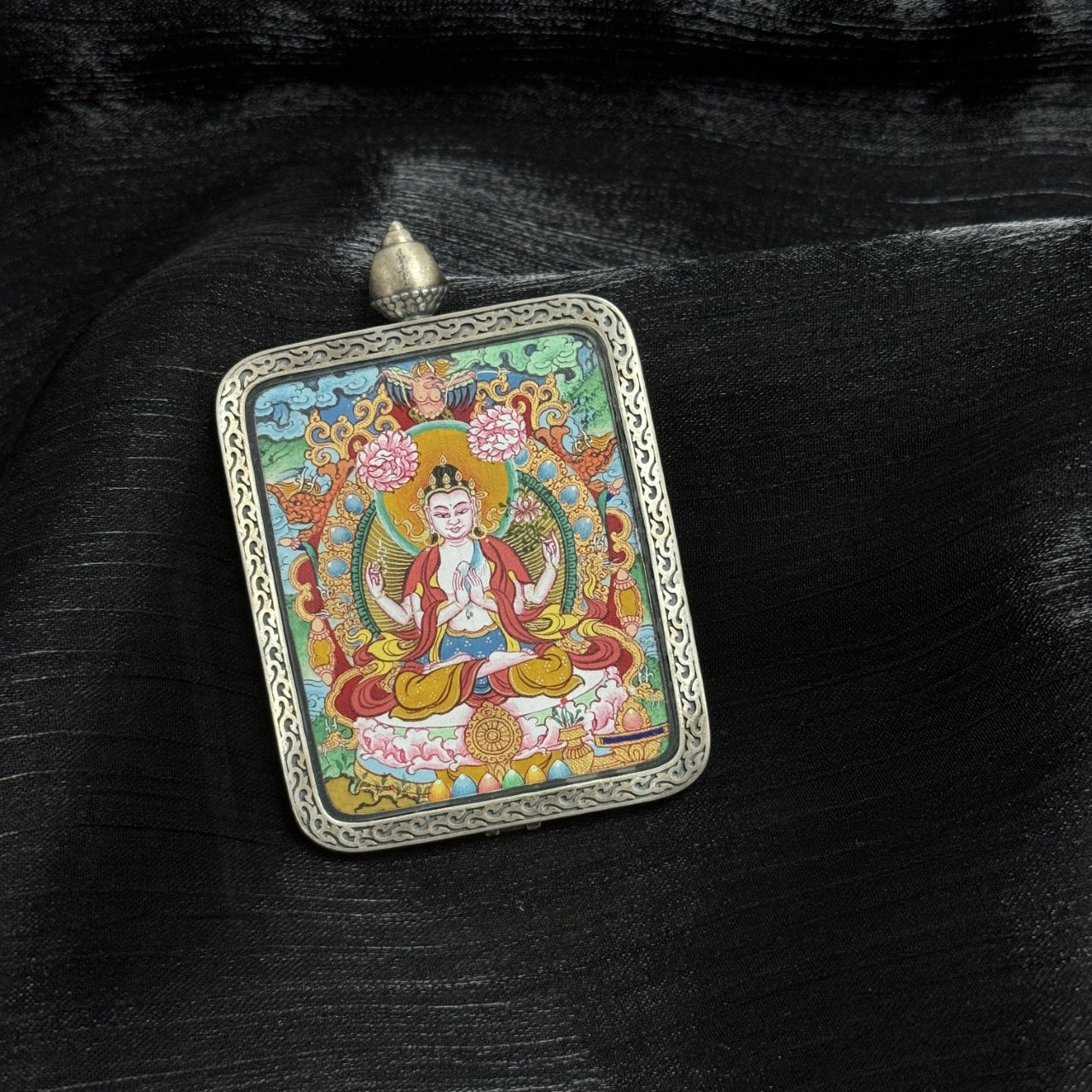 Close-up view of Four-Armed Avalokiteshvara mini thangka pendant showing facial detailing, hand gestures, lotus throne, decorative background motifs, and Dunhuang silver casing texture.